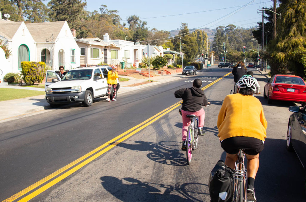 Three bicyclists riding up a straight street, with two women standing outside of their car on the other side of the street, dancing.