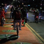 Group of 20 cyclists riding at night on wide road, with center lane painted orange with colorful wheel graphics. The cyclists are taking the right and center lanes. Lights of cars are seen both approaching and going away.