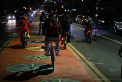 Group of 20 cyclists riding at night on wide road, with center lane painted orange with colorful wheel graphics. The cyclists are taking the right and center lanes. Lights of cars are seen both approaching and going away.