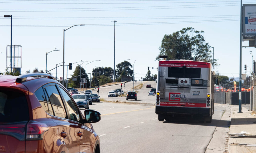 Image of busy multi-lane street with many cars and a bus, heading towards a freeway entrance and overpass. The narrow sidewalk is blocked by poles and debris, and there is no bike lane.