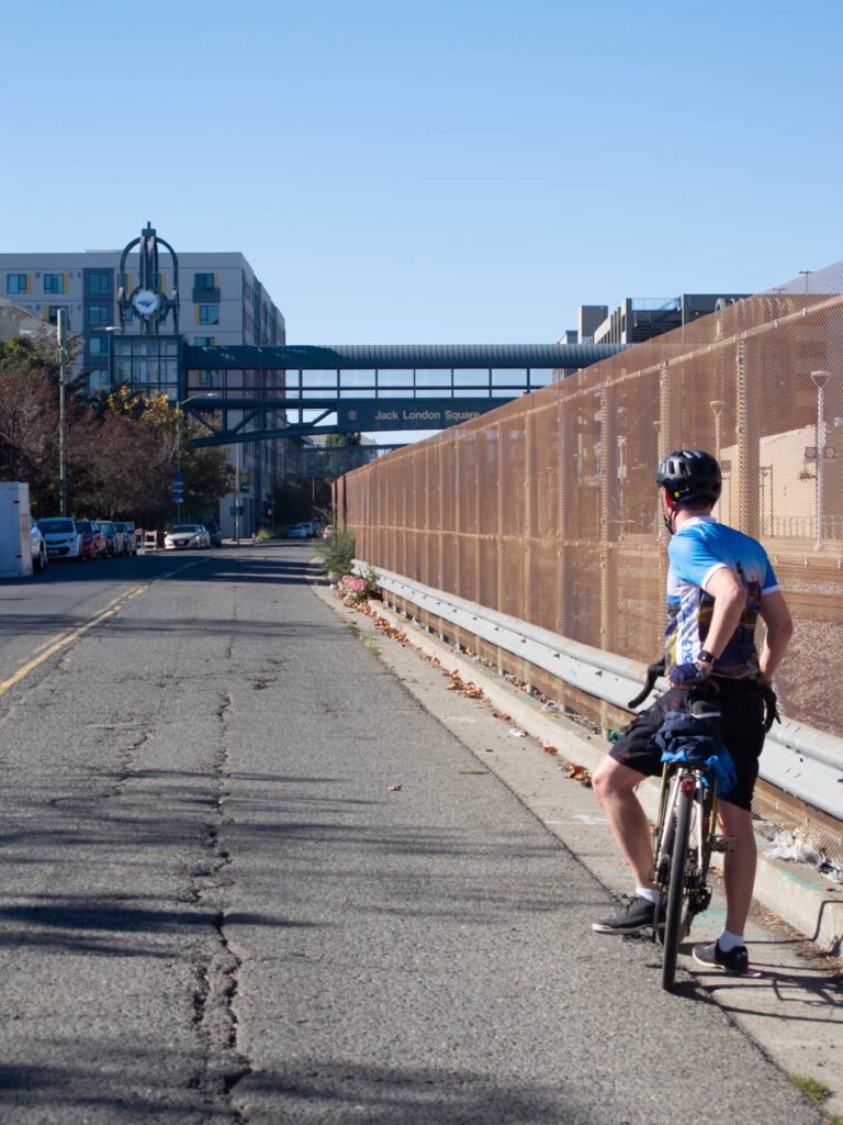 Bicyclist looking down a broken-up road with a high, rusted fence along railroad tracks. A pedestrian bridge over the tracks reads "Jack London Square" 