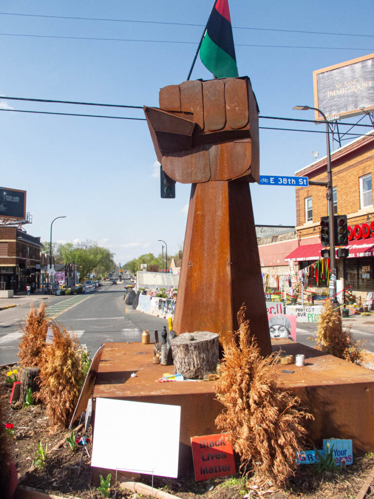 George Floyd Square in Minneapolis, MN. In a road intersection (a sign reads E 38th St.), a central sculpture of rusting iron depicts a raised fist. Atop the fist is a red, black and green flag. At the base are a number of signs, candles and other offerings, along with some plants, mostly dead. The street leading up to the sculpture has a green-painted bike line.