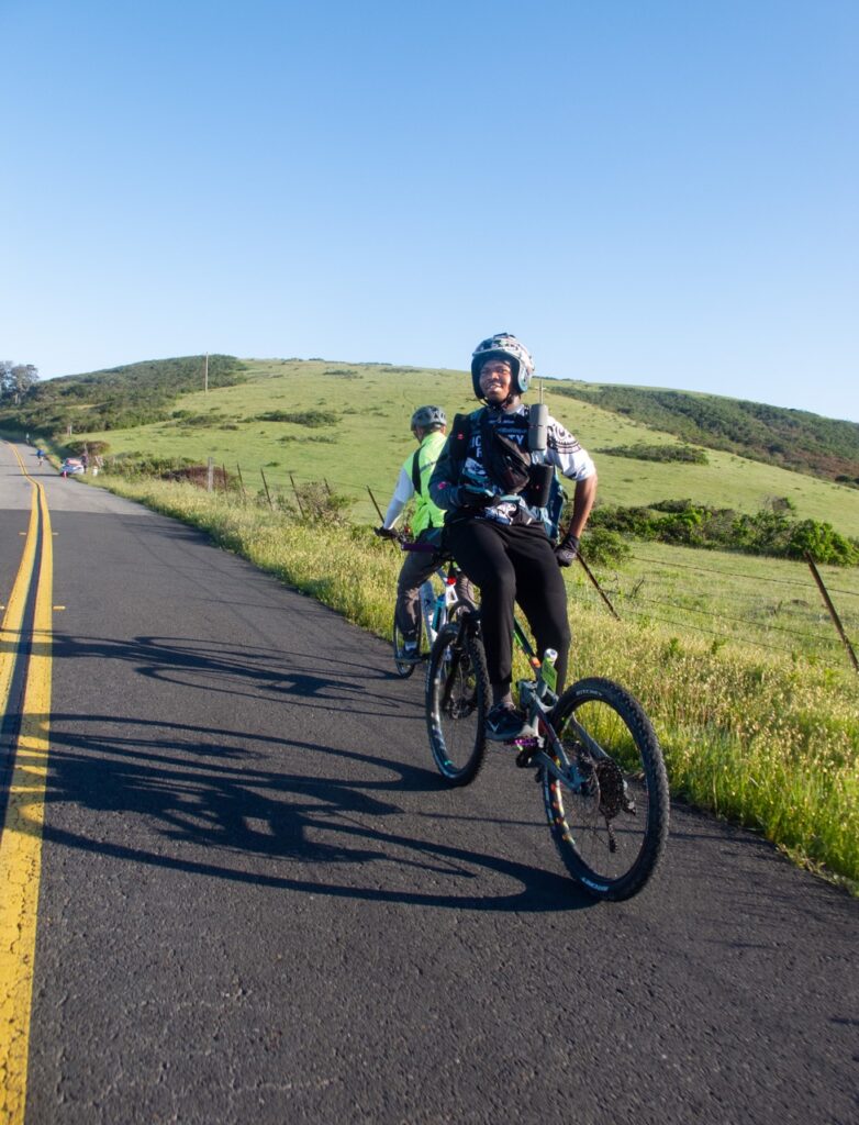Two bicyclists climb a rural road through a grassy hillside on a sunny day in the morning; their shadows are cast across the road. The closer cyclist, a young Black man, is riding his bike backwards, sitting on the handlebars with his feet on the pedals. 