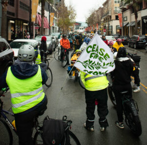 On a rainy day, dozens of bicyclists are standing in one lane of a two-lane commercial street. In the foreground are several people standing with their bikes, wearing yellow safety vests, facing the crowd. One of them has a flag attached to the bike reading, "Cease Fire Now."