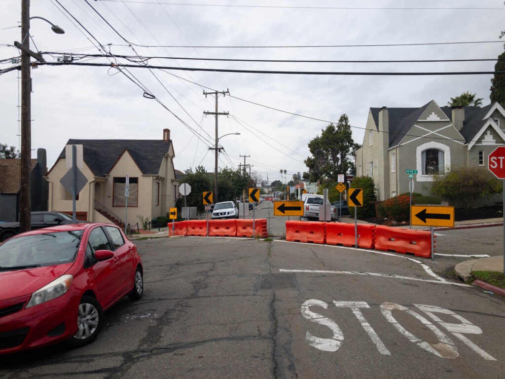 A residential street with somewhat broken-up pavement approaching an intersection with a faded STOP stencil painted in white. The intersection has seven large orange plastic highway barriers installed diagonally across it, with six visible reflective yellow directional signs. The back side of additional signs, presumably directing cars on the other side of the barriers, can also be seen. There is a small gap in the center of the barriers.