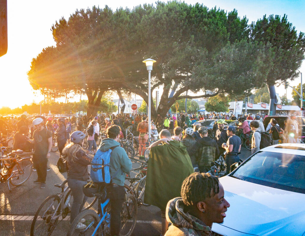 A large crowd of bicyclists gathers in a parking lot. Some appear to be in costume. Rays from a bright sunset enter from the left side of the image. A black man in the foreground of the image smiles as he looks at someone outside of the image on the right side. The canopy of a tree spreads above the group in the center.