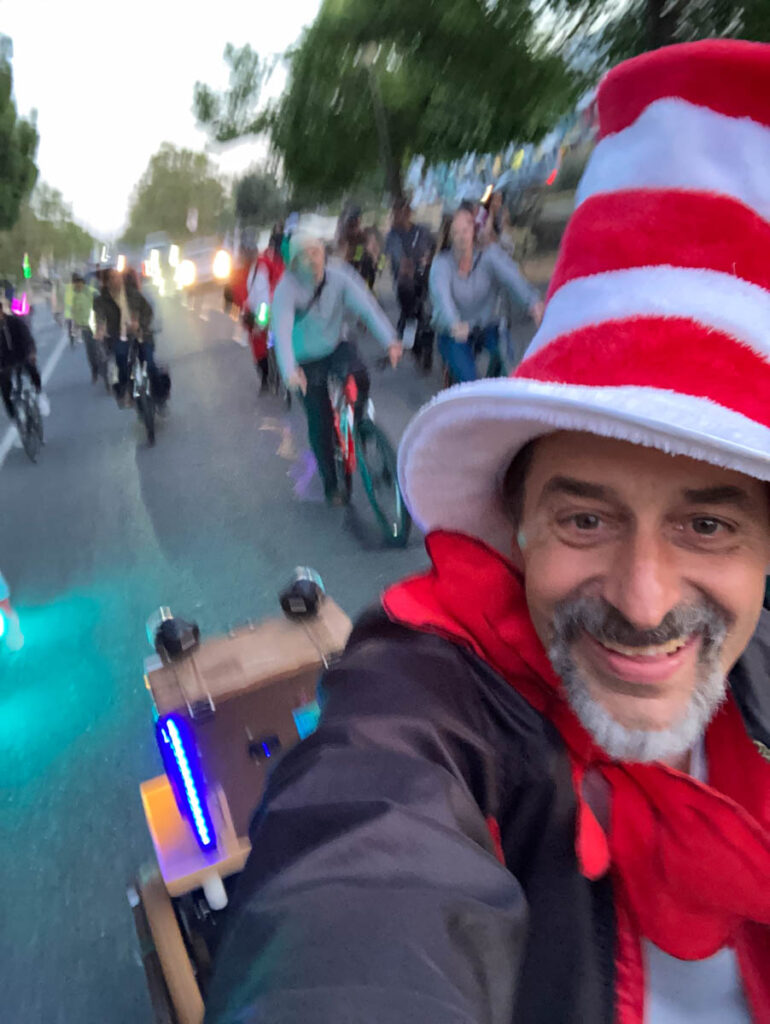 A man takes a selfie while riding a bicycle with a sound trailer decorated with LED strip lights and disco lights. He is wearing a Cat in the Hat costume. He is followed by many cyclists, riding down a wide road with the sunset behind them.