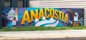 A wide mural on a fence reads "ANACOSTIA". On the left side of the mural are images of Washington, D.C.; the U.S. Capitol, the Washington Monument, and cherry blossoms. A black child holds a hand-made sign with dripping paint reading "I (heart) DC". On the right side of the mural, three Black musicians play (a guitarist, trombone player, and keyboard)
