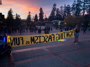 At UC Berkeley's Sproul Plaza, under a sunset sky with pink clouds, a photograph shot from behind a woman holding up a yellow banner reading "Fighting Fascism is Fun". Some people are taking pictures of the banner; others are milling about on the plaza.