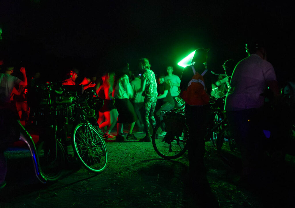 A crowd of people at night, lit by green and red LED lights including a triangular green flag, dancing among many bicycles.