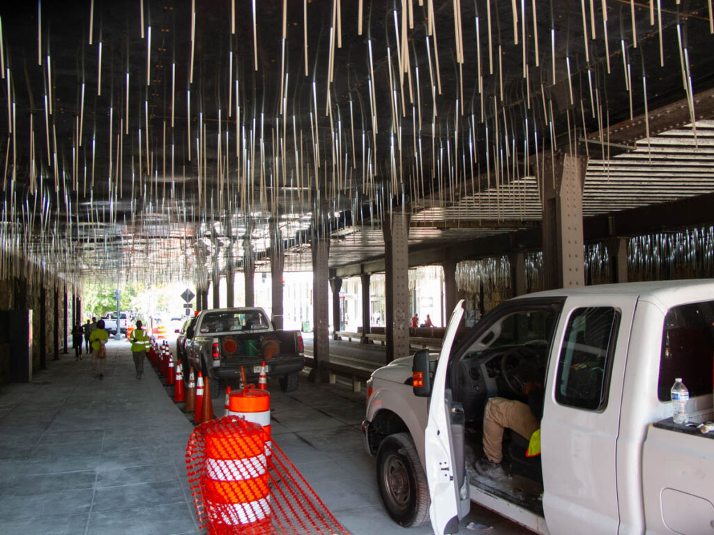 A construction project in an underpass. Sparkly lights hang from the ceiling. Three pickup trucks are parked in what appears to be a bike lane under construction. Orange cones line the bike lane. Construction workers in reflective vests are seen.
