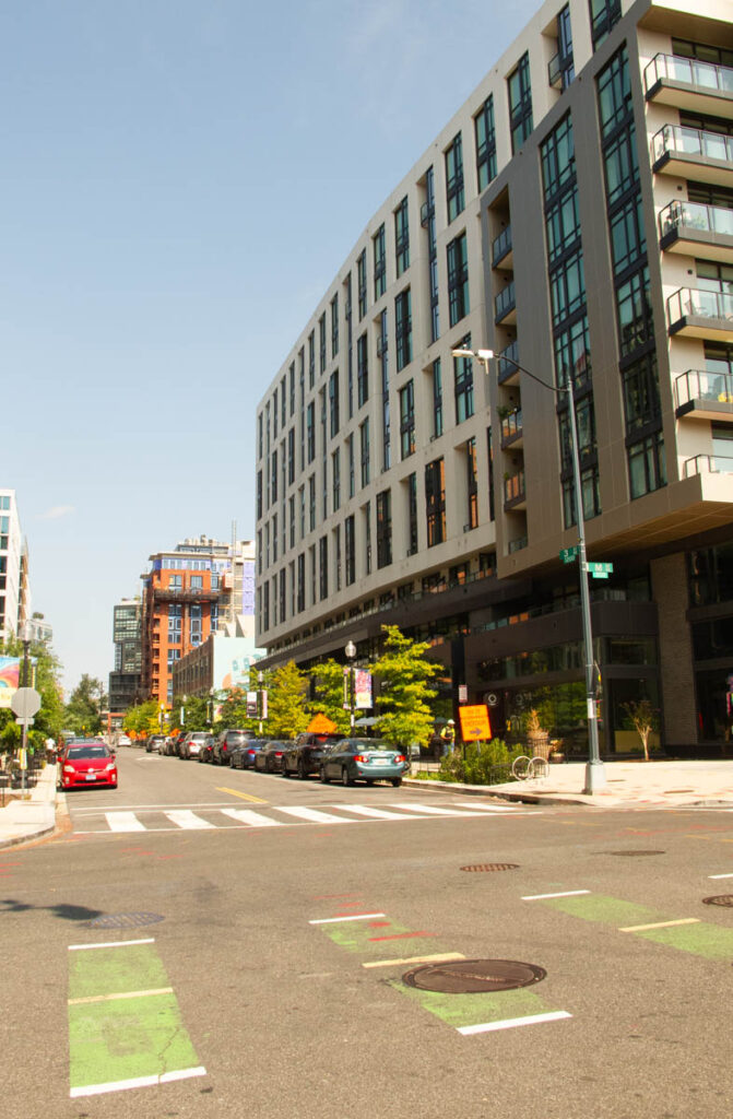 A streetscape of cars parked along a fairly newly-landscaped street, with recently built 5-story apartment buildings on both sides. Orange construction detour signs are seen on the far sidewalk.