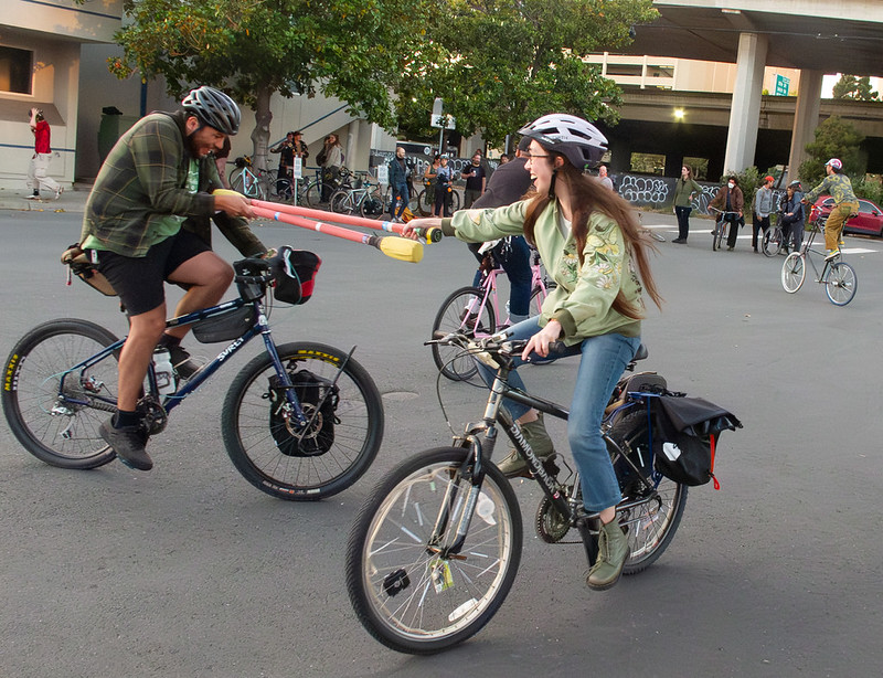 In an urban alley, two laughing people on bicycles ride towards each other with lances made of pool noodles. A crowd watches in the background.
